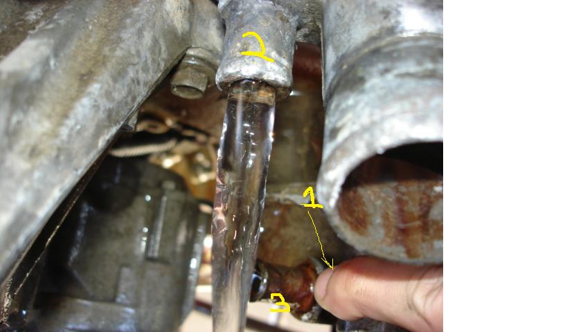 Close-up of leaking metal coolant tube and corroded fitting under an engine, with yellow numbered annotations