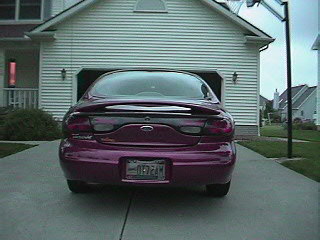 1999 Ford Taurus - Rear view of a purple 1999 Ford Taurus with a spoiler, parked in front of a house and garage.