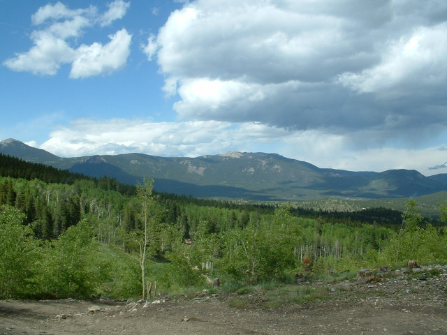 Mountain landscape with green forested hills and cloudy blue sky, dirt ground in foreground