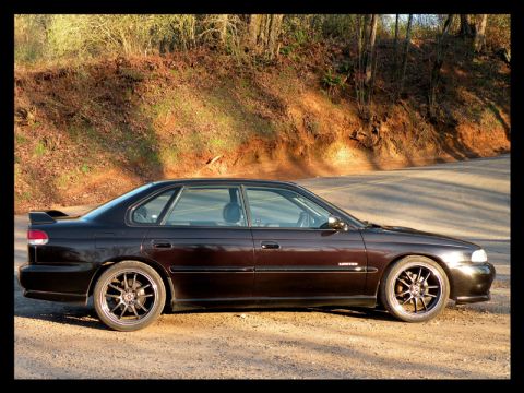 1998 Subaru Legacy - This dark sedan, possibly a 1998 Subaru Legacy, is parked on a dirt road with a wooded hillside behind it.