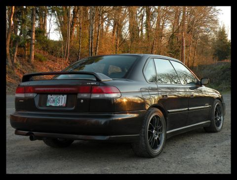 1998 Subaru Legacy - The rear of a black Subaru Legacy GT sedan is shown with a spoiler and aftermarket wheels.