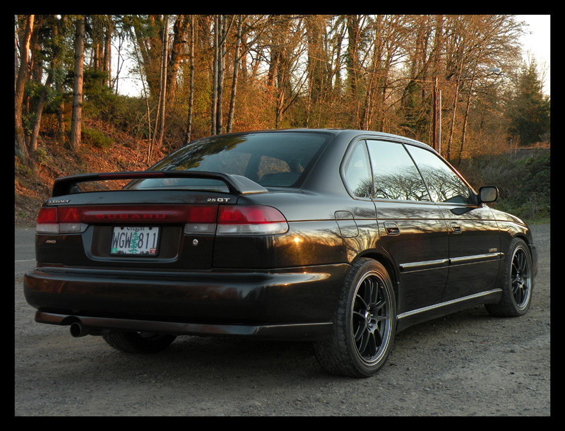 Black Subaru Legacy GT sedan, rear 3/4 view, parked on dirt road with trees in background