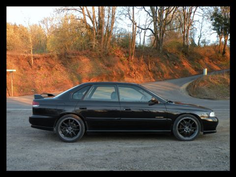 1998 Subaru Legacy - A black 1998 Subaru Legacy sedan is viewed from the side, parked on a gravel lot with a wooded hillside in the background.