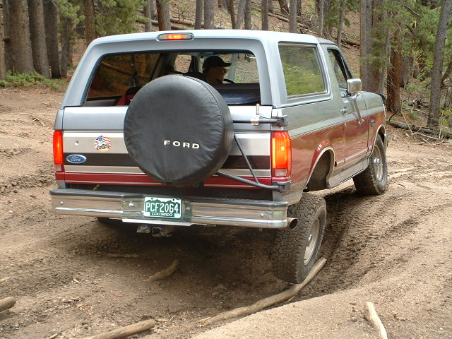 Rear view of a lifted two-tone silver/red Ford Bronco on a rutted forest dirt trail, black FORD spare tire cover on tailgate.
