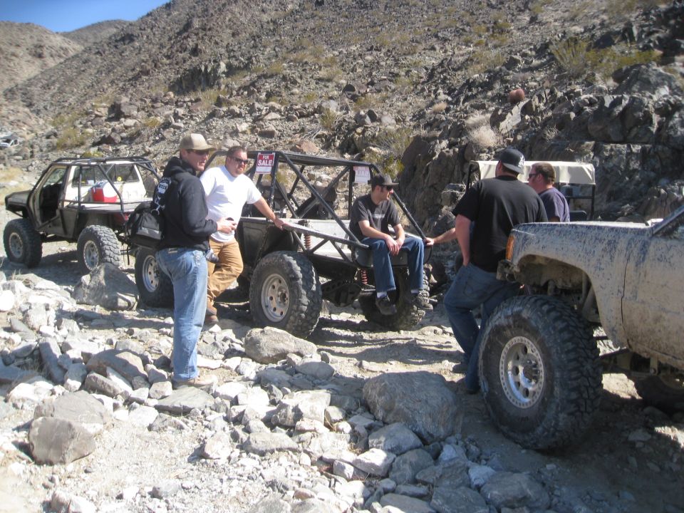 Several modified off-road vehicles and people gathered on a rocky, desert-like terrain. One vehicle has a 'FOR SALE' sign