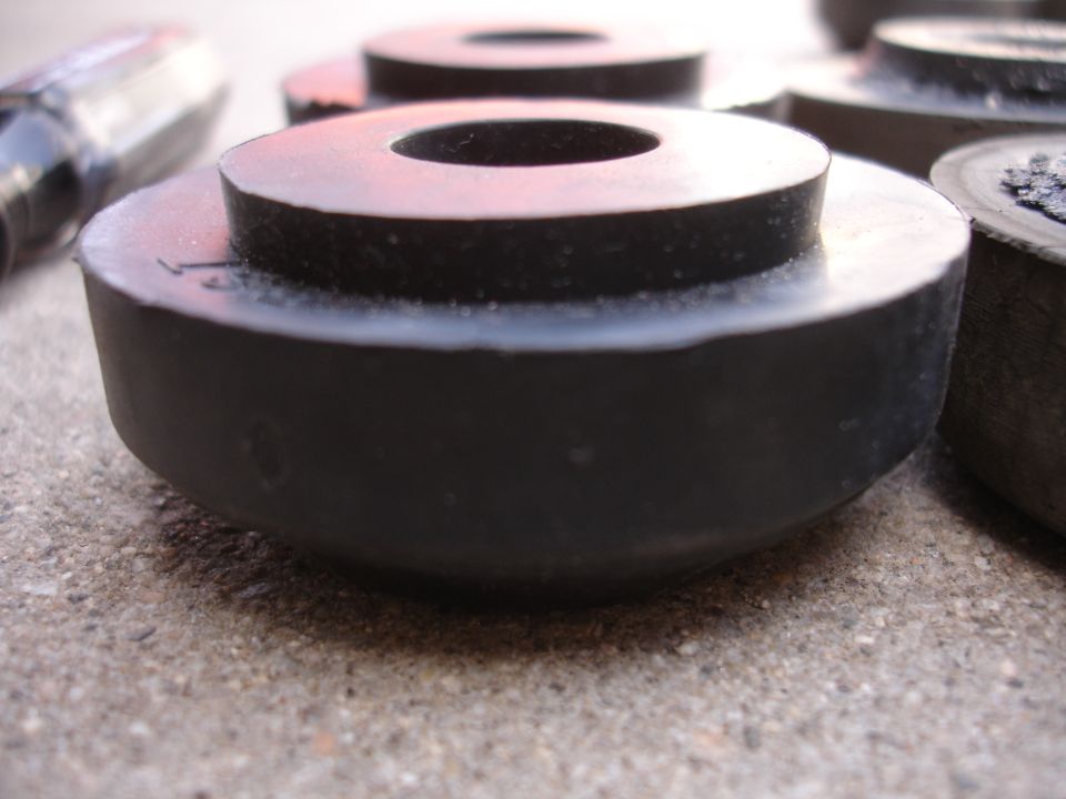 Close-up of several black rubber suspension bushings on a concrete surface. A tool handle is visible on the left