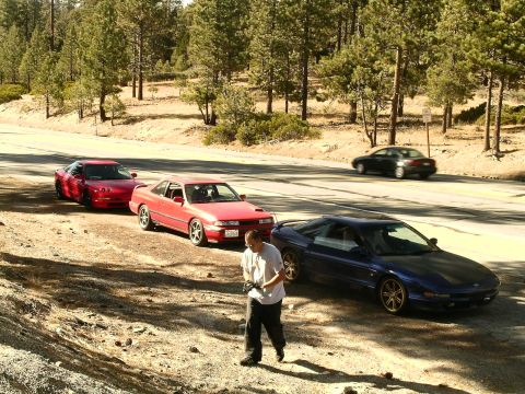1993 Ford Probe - A group of sports coupes are parked on the side of a road in a wooded area, with a person standing near one of them.