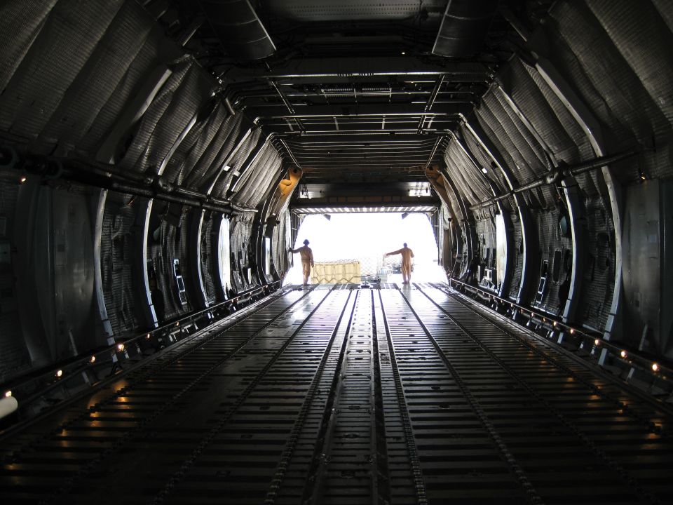 Inside cargo plane looking toward open rear ramp, two people visible near the exit