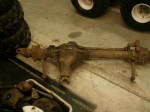 1990 Ford Bronco - A bare, rust-coated solid front axle housing lying on a concrete shop floor, with the offset cast center section, differential cover area,…