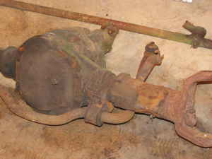 1990 Ford Bronco - A rusty solid front axle assembly sitting on a dirt floor, showing the cast differential housing, coil spring perch, radius arm bracket and…