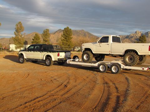 1995 Ford Bronco - A white pickup truck is being towed by another pickup truck on a trailer in a dry, dusty area.