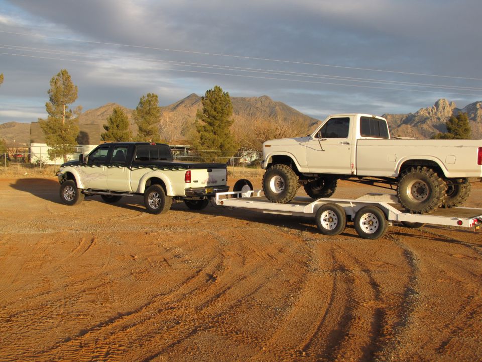 White pickup truck on trailer being towed by another white pickup truck. Mountains in background