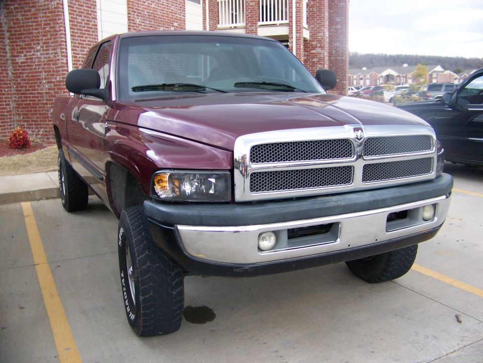 Maroon Dodge Ram pickup with chrome grille and off-road tires parked by a brick building