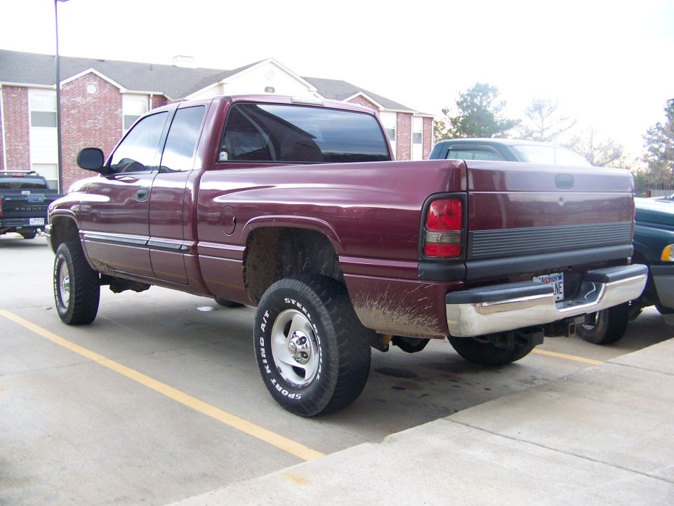 Maroon extended-cab pickup with muddy rear fender and white-letter tires, rear driver-side view in parking lot