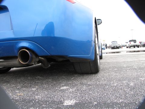 2003 Mazda Protege - Rear view of a blue car with a prominent exhaust pipe and wide tire, parked outdoors with other vehicles.