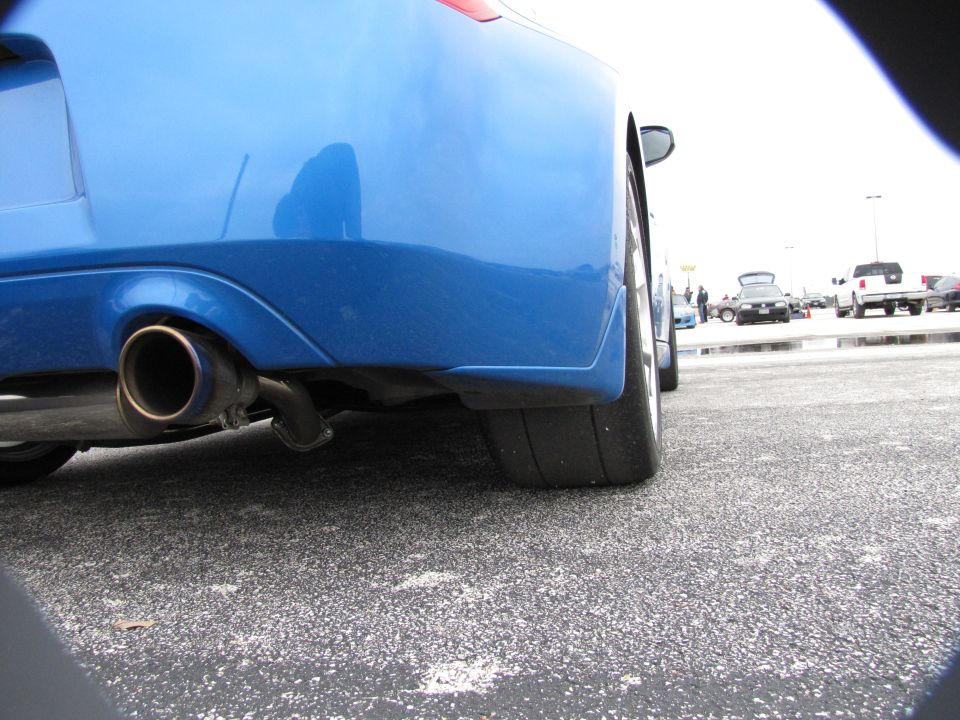 Close-up of a blue car's rear bumper, showing the exhaust pipe and a wide tire. Other vehicles are visible in the background