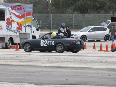 2003 Mazda Protege - A dark convertible sports car with racing numbers is parked on a track next to a trailer and another vehicle.