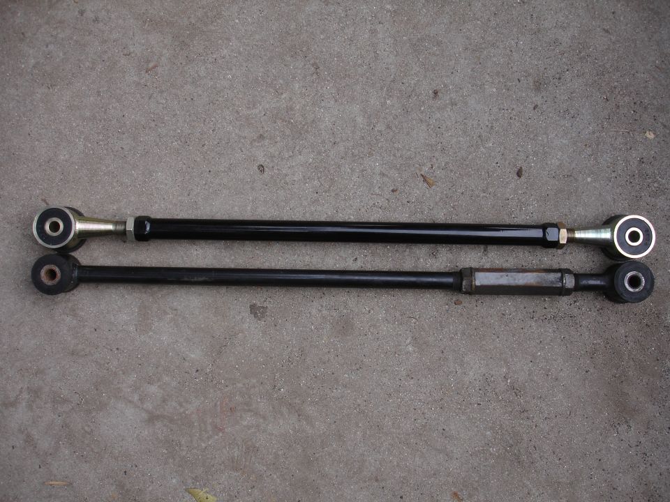 Two black suspension control arms with metal and rubber bushings on a concrete surface