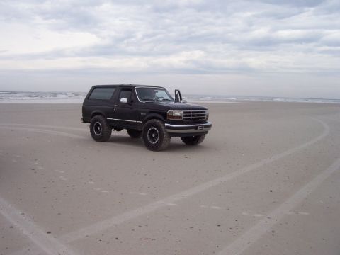 1994 Ford Bronco - A black 1994 Ford Bronco is parked on a sandy beach with the ocean in the background under a cloudy sky.