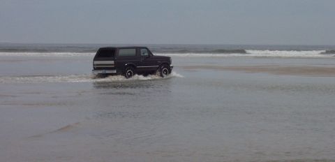 1994 Ford Bronco - A dark 1994 Ford Bronco navigates shallow water and waves on a sandy beach.