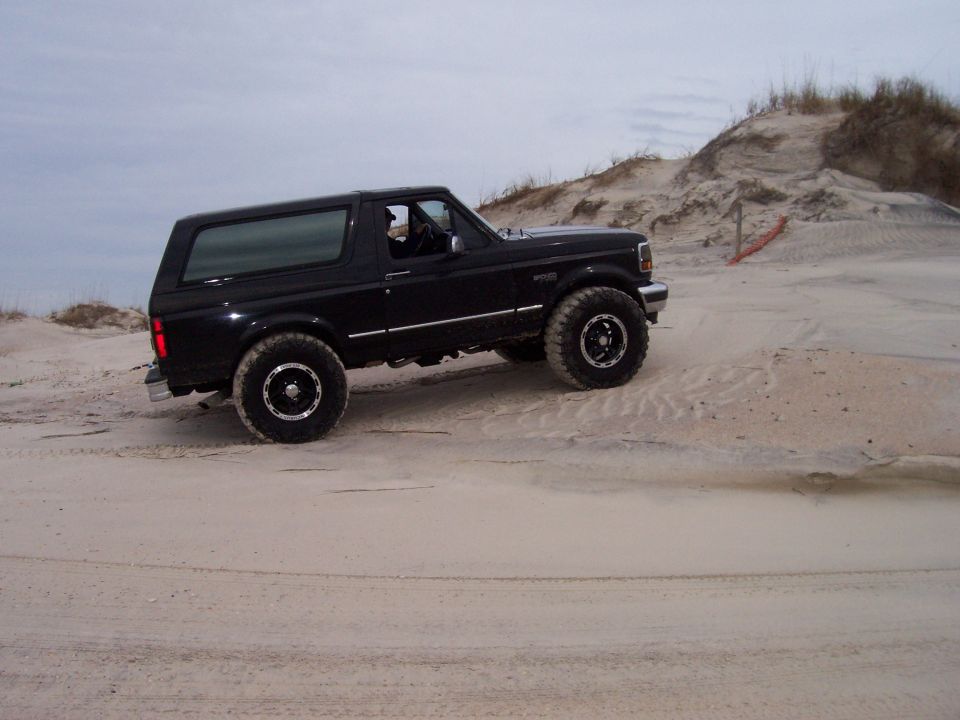 Black 1994 Ford Bronco driving on a sandy beach with dunes in the background