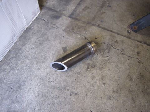1996 Ford Bronco - A single metal exhaust tip rests on a cracked concrete surface, with a white corrugated object in the background.