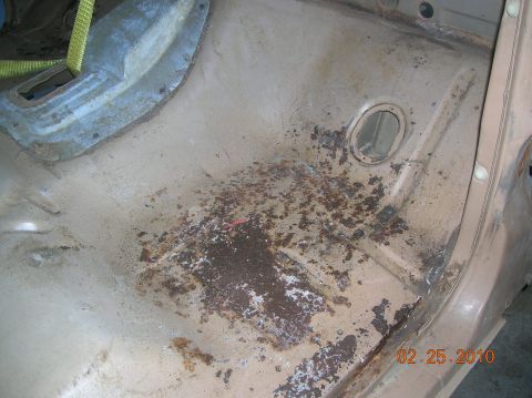 1985 Ford Bronco - The interior floor of a vehicle shows significant rust and wear, with a yellow strap visible above.