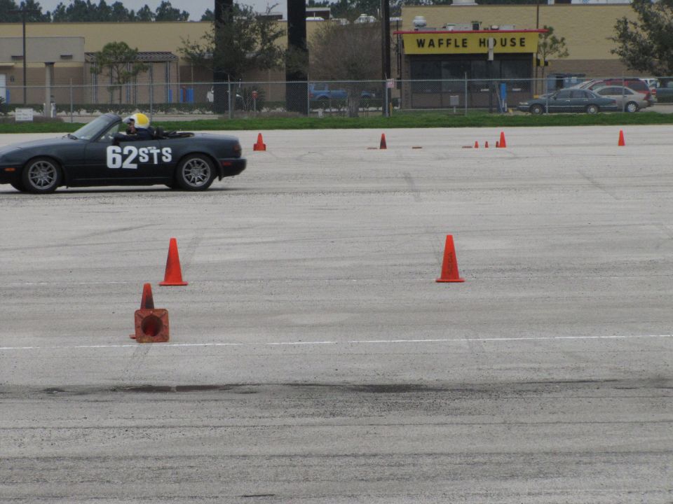 Black convertible sports car with white racing numbers '62STS' on the side, driving on a track with cones