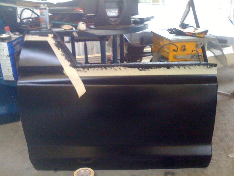 Black car door with masking tape on the top edge, in a workshop setting with tools visible