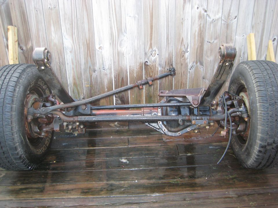 Removed solid front axle with wheels and steering linkage sitting on a wet wooden deck