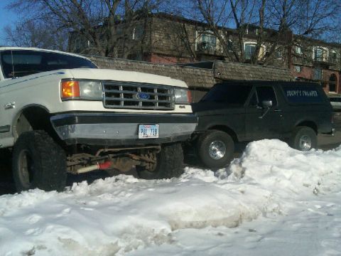 1987 Ford Bronco - Two Ford trucks, a Bronco and a larger pickup, are parked in the snow. The Bronco has text on its side.