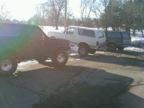 1987 Ford Bronco - Three SUVs are parked in a lot on a sunny day with snow on the ground.
