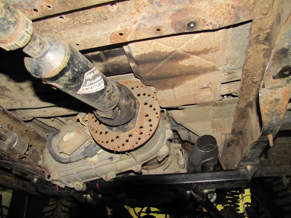 Underside of a vehicle showing a driveshaft, transfer case, and exhaust pipe. A label on the driveshaft reads 'High Impact Drivelines'
