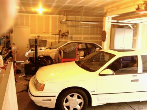 1989 Ford Taurus - A white 1989 Ford Taurus sedan with a sunroof is parked in a garage next to another vehicle.