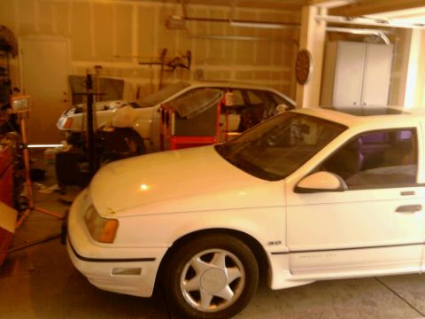 1989 Ford Taurus - A white sedan with a sunroof is parked in a garage, with another vehicle visible behind it.