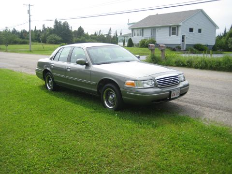 1999 Ford Crown Victoria - A gray Ford Crown Victoria is parked on a grassy area next to a road, with a house in the background.