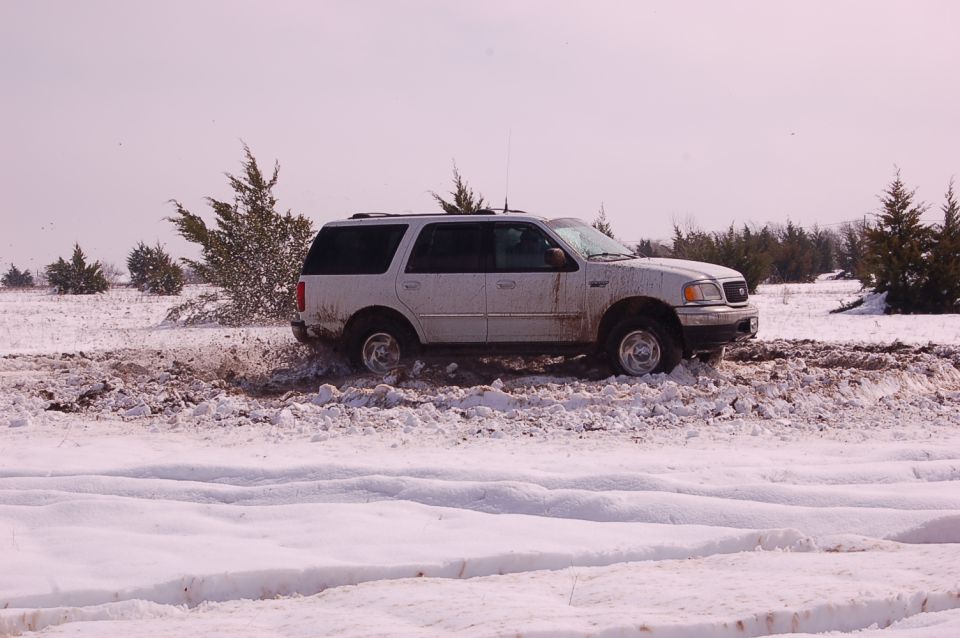 White SUV stuck in snow, tires spinning in mud and snow. Trees and open field in background