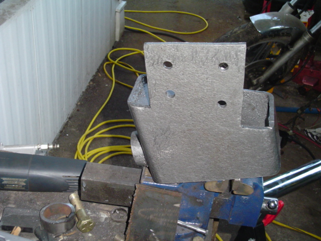 Bare steel fabricated mounting bracket with four bolt holes clamped in a blue bench vise