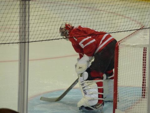 1996 Ford Bronco - A hockey goalie in a red uniform is positioned in front of the net, bent over with their stick on the ice.