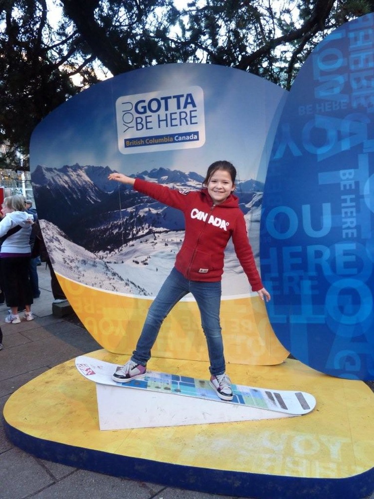 Girl in red hoodie and jeans on snowboard, standing on a yellow platform in front of a large blue display with mountains and text