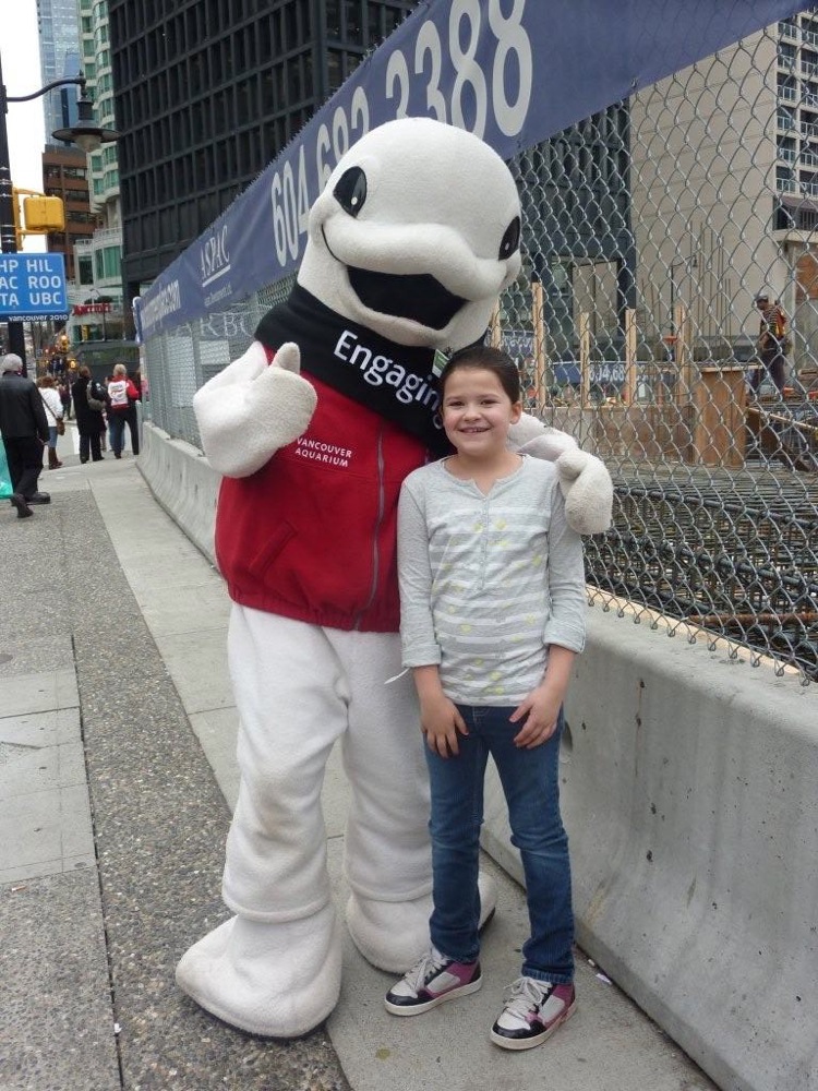 Mascot in red vest and white suit poses with a child on sidewalk. Banner behind them has phone number