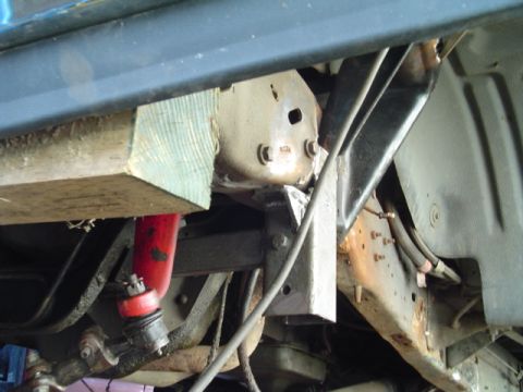 1993 Ford Bronco - Close-up under a raised body showing the frame rail and a body-mount bracket with a wooden block wedged between the frame and the floor pan…