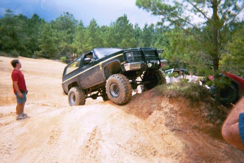 1985 Ford Bronco - A lifted Chevrolet K5 Blazer navigates a steep dirt incline with trees in the background.