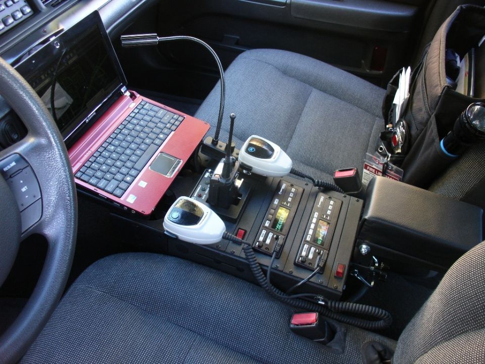 Interior of a police car showing a laptop, two-way radios, and control panels between the seats