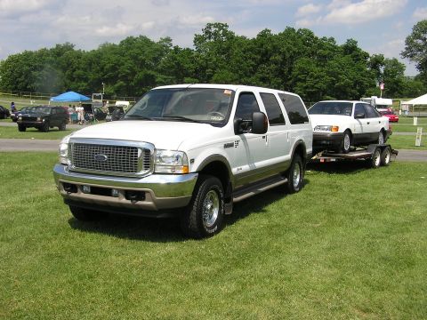 2000 Ford Excursion - A white 2000 Ford Excursion is shown from the front 3/4 angle, parked on a grassy field and towing a white sedan on a trailer.
