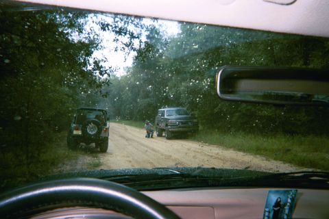 1985 Ford Bronco - Two SUVs navigate a dirt road surrounded by trees, with one vehicle stopped and a person kneeling beside it.