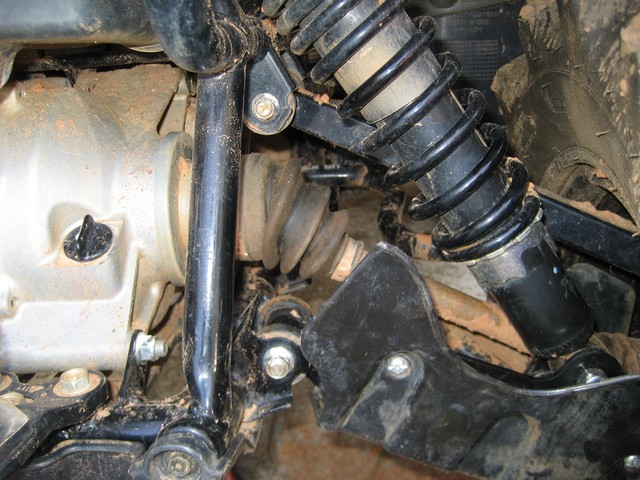 ATV suspension with black shock absorber, drive shaft boot, and metal frame components. Mud is visible