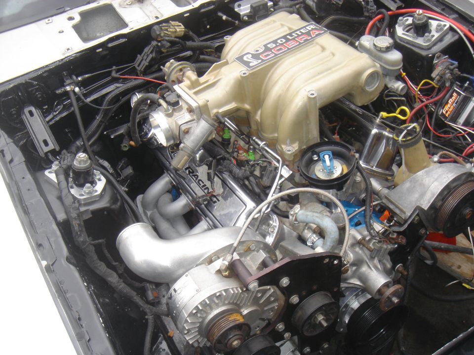 Ford 5.0 Cobra engine with supercharger and custom exhaust headers. "RACING" is visible on valve cover