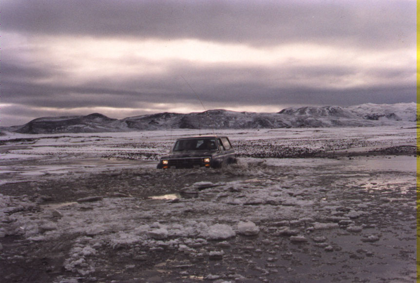 Dark vehicle crossing snowy flats with distant hills under cloudy sky