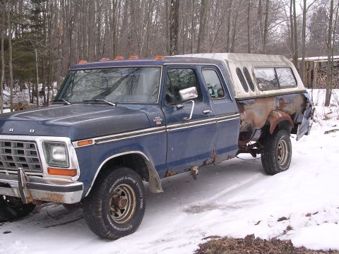 1979 Ford F-150 - A blue Ford pickup truck with a camper shell sits in a snowy, wooded area. The rear of the truck shows significant rust damage.